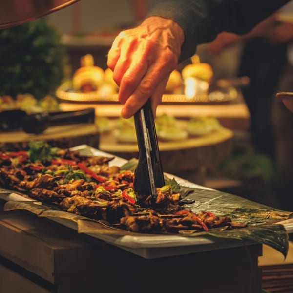 A hand uses tongs to serve skewers of grilled meat and vegetables from a buffet.