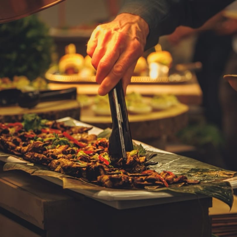 A hand uses tongs to serve skewers of grilled meat and vegetables from a buffet.