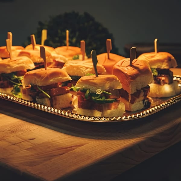A silver tray filled with numerous mini sliders, each with a toothpick, on a wooden surface.