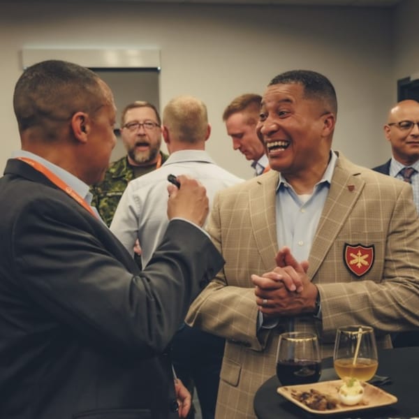 Two men in suits talking and laughing, with drinks and appetizers on a table in the foreground.