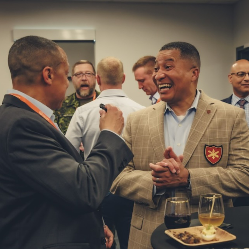 Two men in suits talking and laughing, with drinks and appetizers on a table in the foreground.