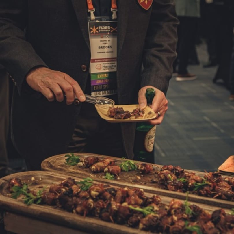 A person in a suit uses tongs to serve bacon-wrapped appetizers from a wooden tray.