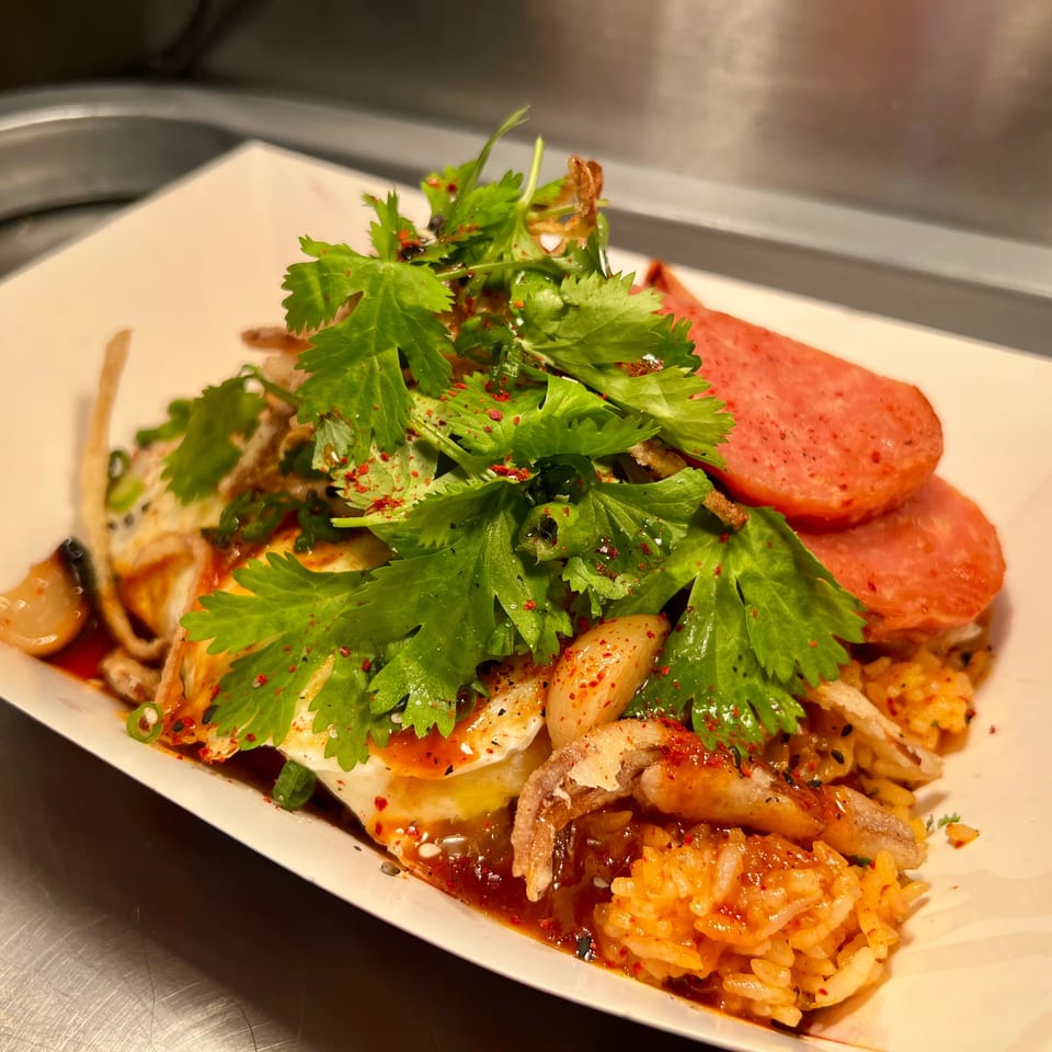 Spicy rice dish with fried eggs, sliced meat, cilantro, and chili flakes in a white container.