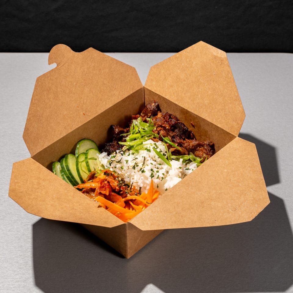 Open cardboard takeout box filled with rice, meat, cucumbers, carrots, and kimchi.