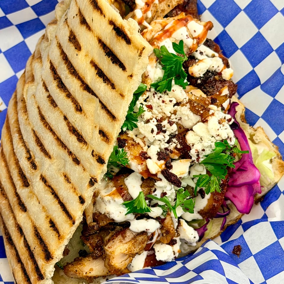 Grilled flatbread sandwich filled with chicken, feta, parsley, and purple cabbage on checkered paper.