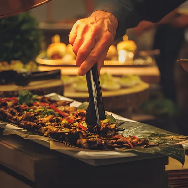 A hand uses tongs to serve skewers of grilled meat and vegetables from a buffet.