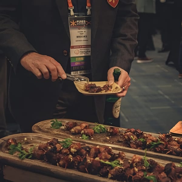 A person in a suit uses tongs to serve bacon-wrapped appetizers from a wooden tray.