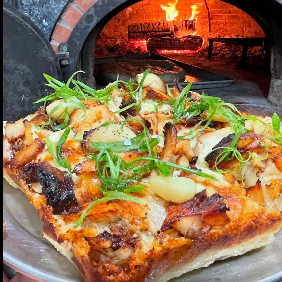 A square pizza with chicken, garlic, and green onions, in front of a wood-fired oven.