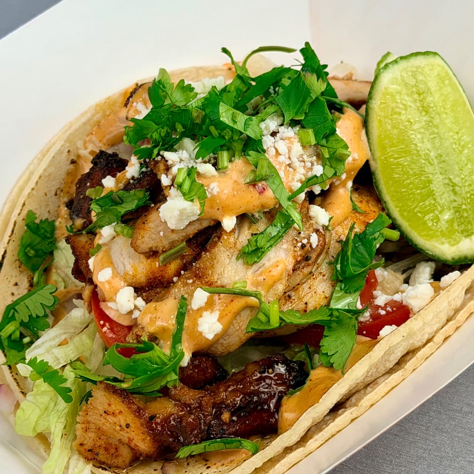 Close-up of a taco with grilled chicken, sauce, cilantro, and feta cheese, served with a lime wedge.