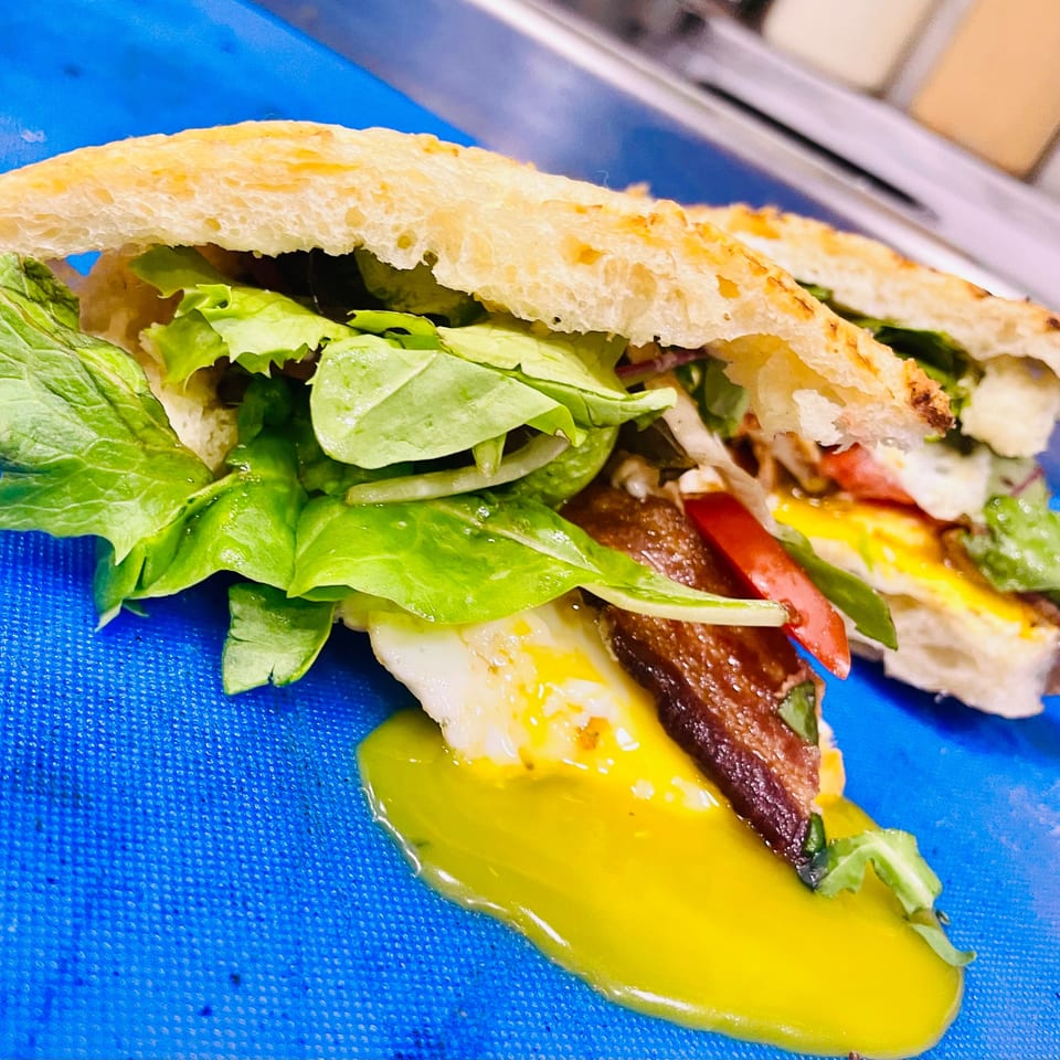 A Little Bear Street Eats sandwich with bacon, egg, lettuce, and tomato on a blue surface.