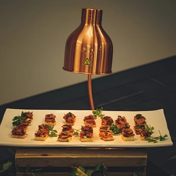 A white platter of appetizers with meat and parsley under a copper heat lamp.